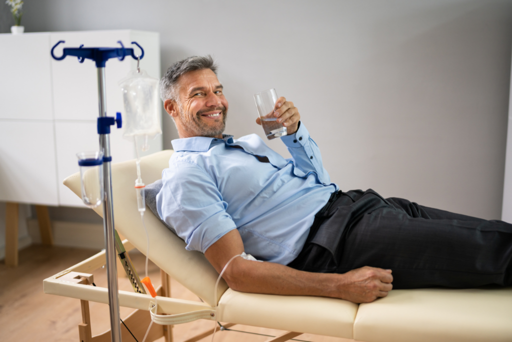 Middle-aged man receiving Bioidentical Hormone Therapy treatment in a clinic, smiling and holding a glass of water while relaxing on a treatment chair.